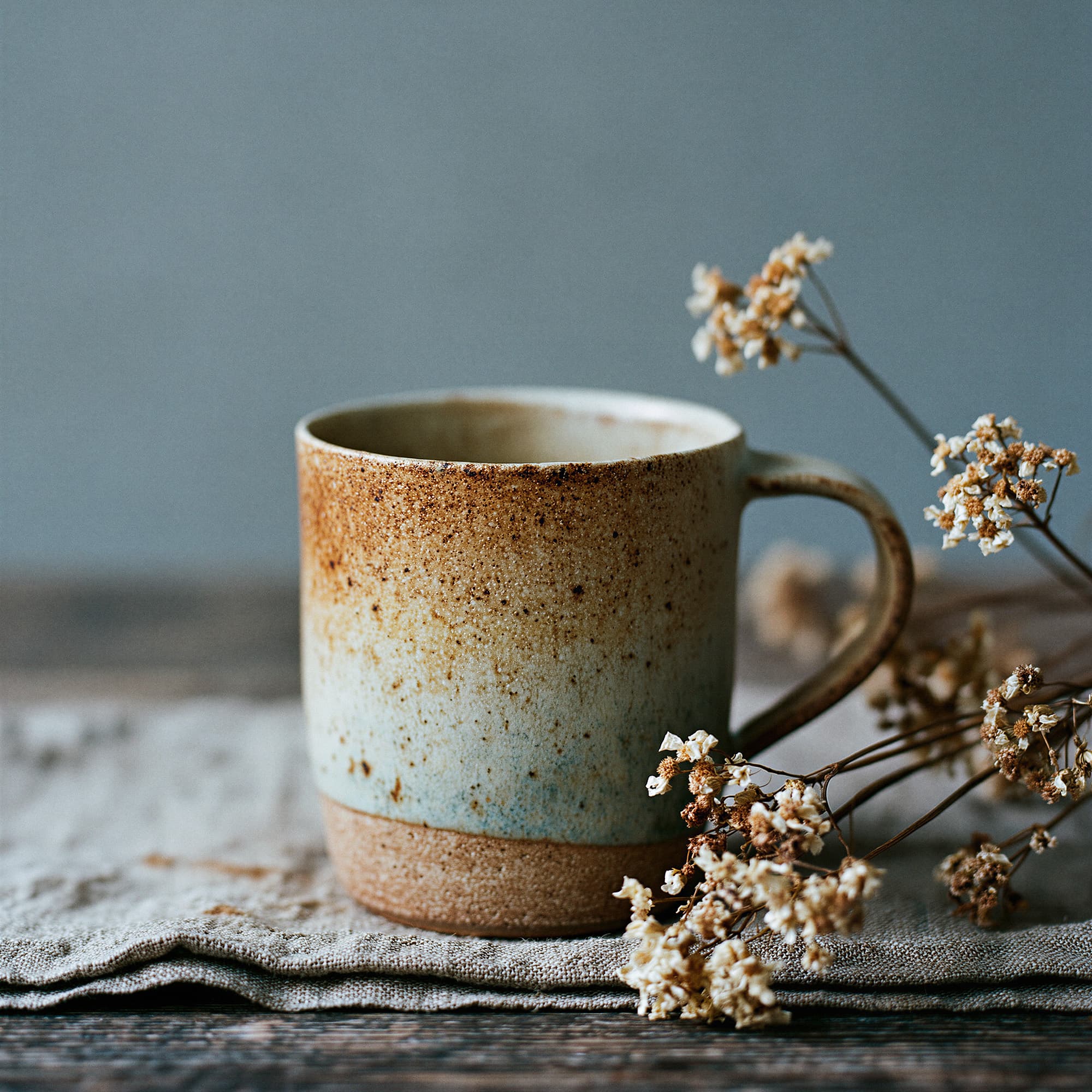 Hand-thrown stoneware drinking mug with iron oxide speckle and celadon glaze on aged linen with dried botanicals, simulated product photography created with Midjourney, stamped with VISID structured metadata